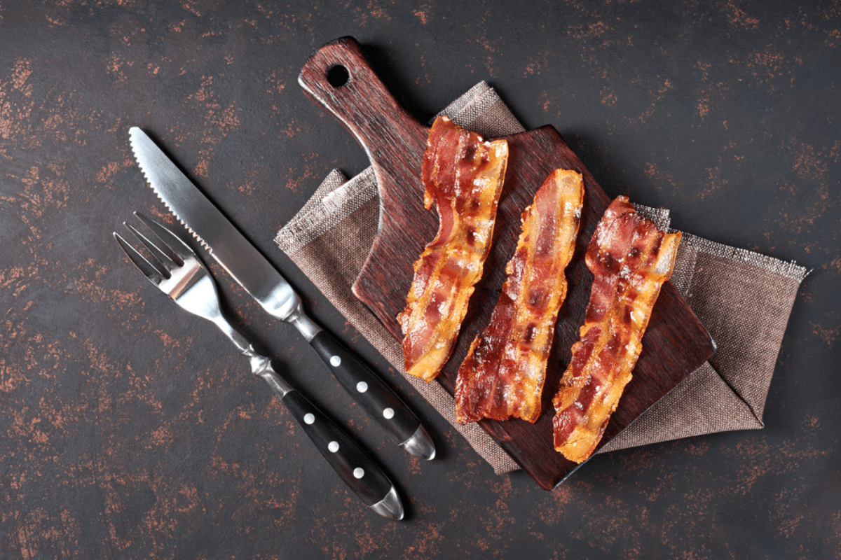 Tre fette di pancetta poste su un tritatutto in legno insieme a coltello e forchetta